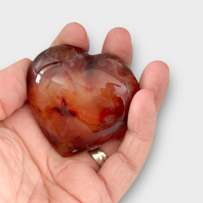 A polished Carnelian Heart resting in the palm of a hand, highlighting its compact size, vibrant orange color, and smooth, heart-shaped design.